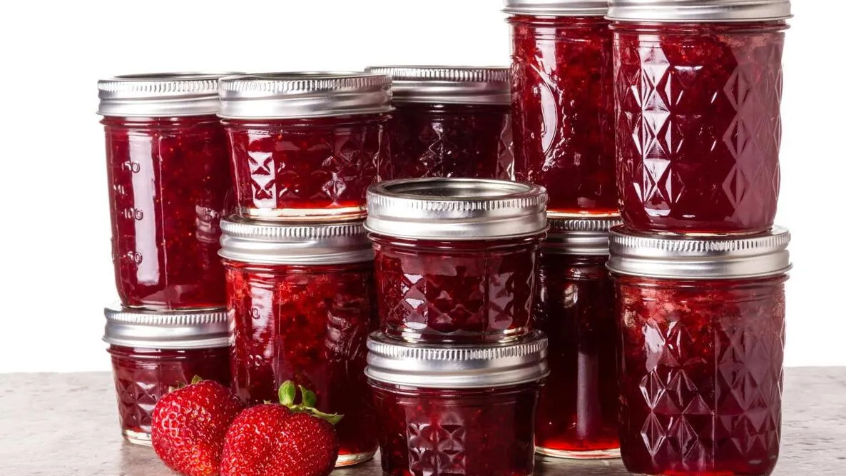 A stack of strawberry preserves, in glass canning jars of different sizes. There are also two ripe strawberries, for added visual appeal.
